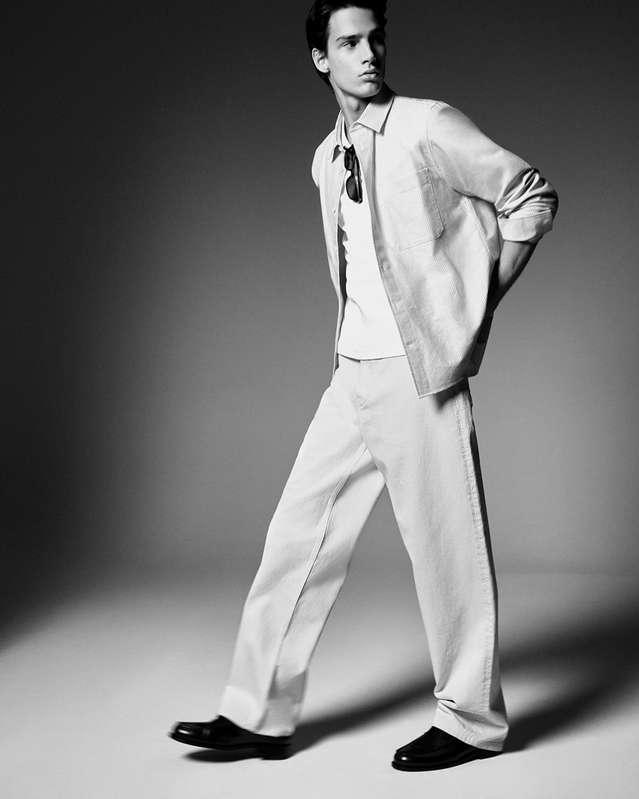 Black and white photo of a person in a casual outfit, wearing a light shirt, white t-shirt, and trousers, walking confidently.