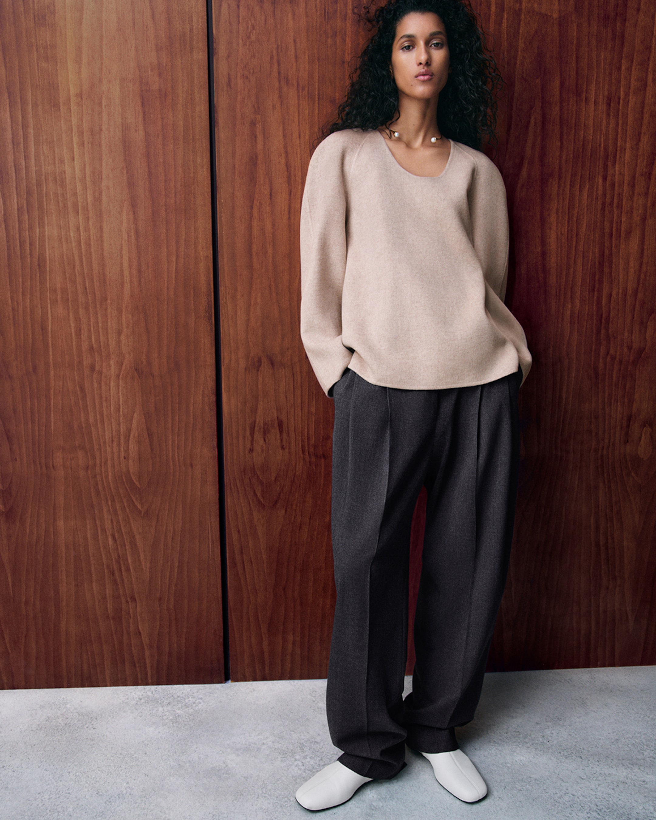 Person in a beige sweater and dark pants leans against a wooden wall, wearing white shoes and a necklace, with a confident expression.