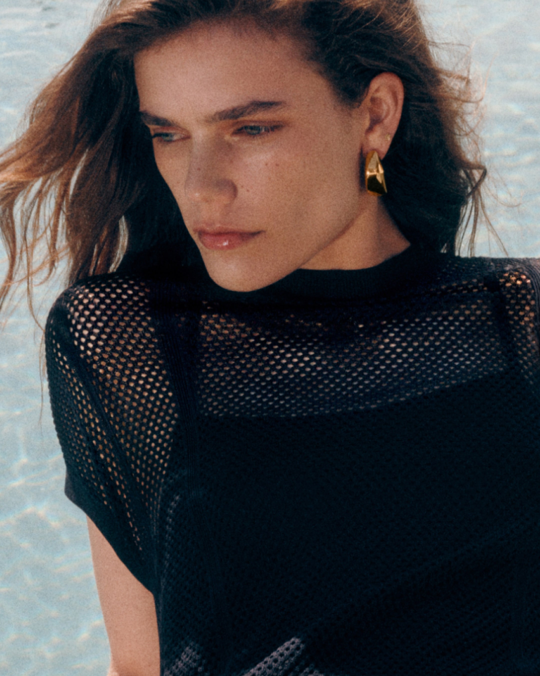 Woman with long hair wearing a black mesh top and gold triangular earrings, gazing downward against a light blue background.
