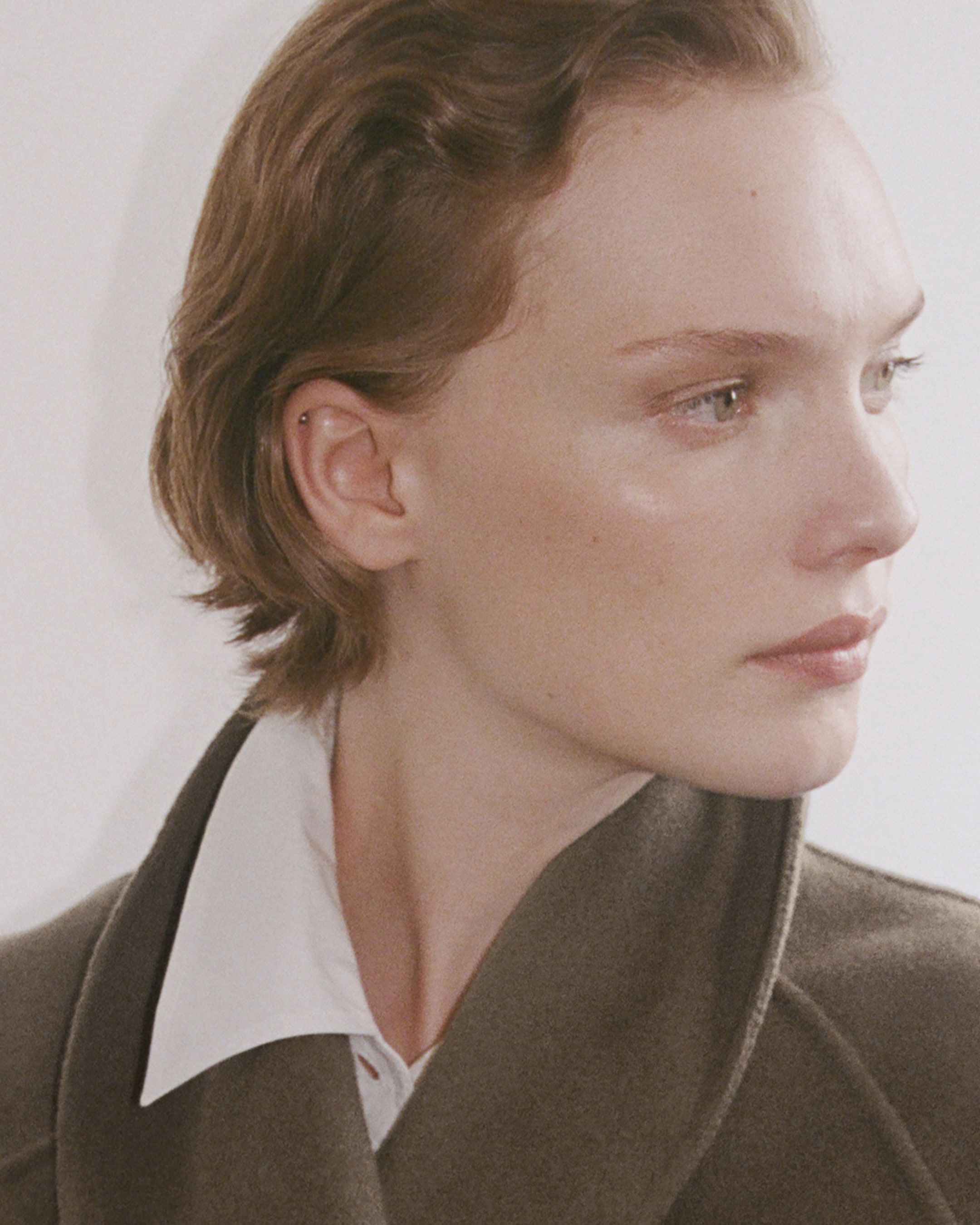 Person with short hair wearing a white shirt and brown coat, gazing to the side with a neutral expression.