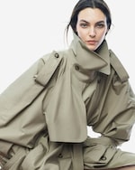 Placeholder for: Person wearing an oversized beige trench coat with high collar, looking directly at the camera against a plain white background.