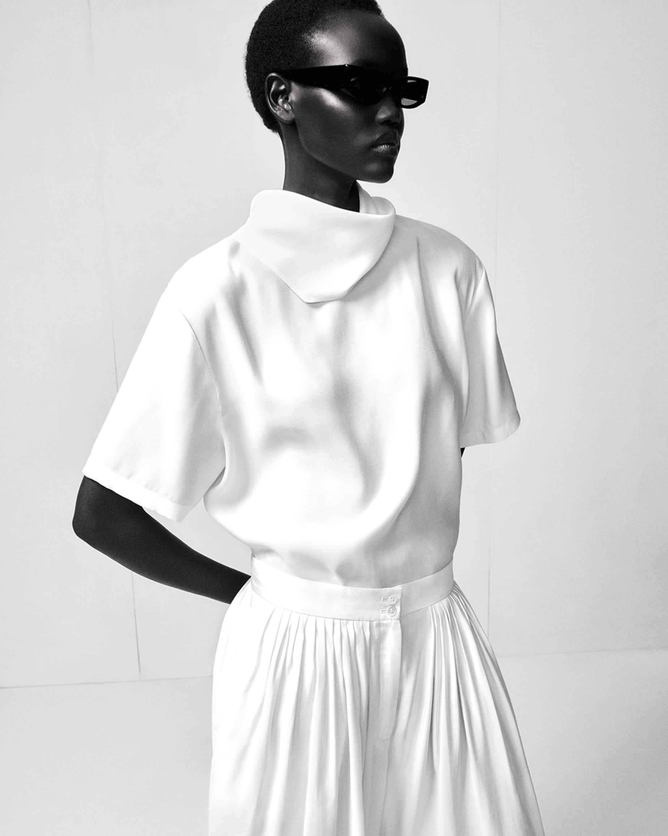 A person in a stylish white outfit with a high collar is wearing dark sunglasses, posing confidently in a monochromatic setting.