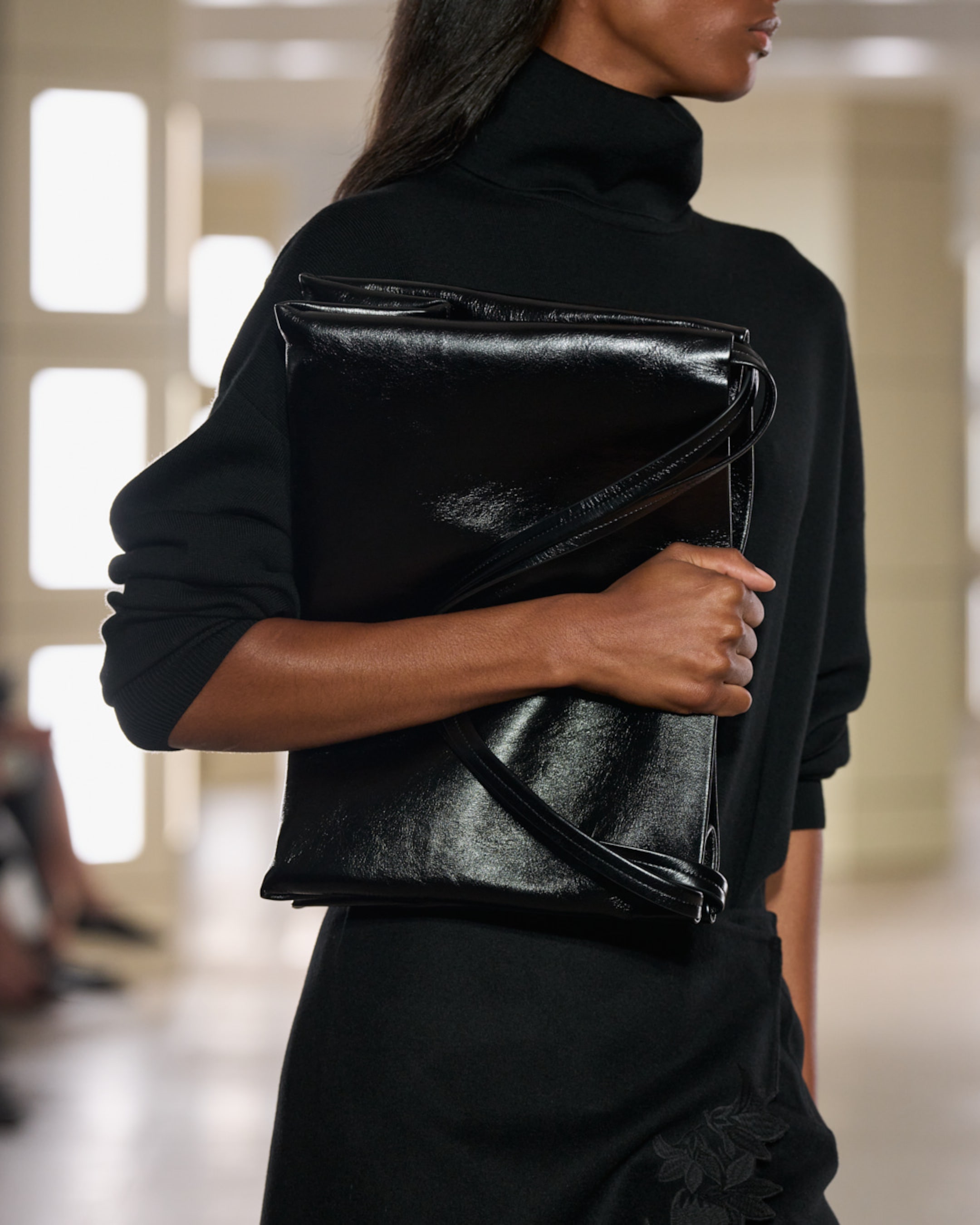 A person in a black high-neck sweater holds a shiny black leather clutch, standing indoors with a blurred background.