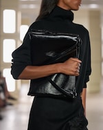 Placeholder for: A person in a black high-neck sweater holds a shiny black leather clutch, standing indoors with a blurred background.