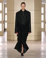 Placeholder for: Model wearing a chic, oversized black coat and loafers walks down a minimalist runway with beige paneling and soft lighting.
