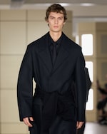 Placeholder for: A person with short hair wears a dark, oversized suit and walks on a runway, with a sweater tied around their waist.