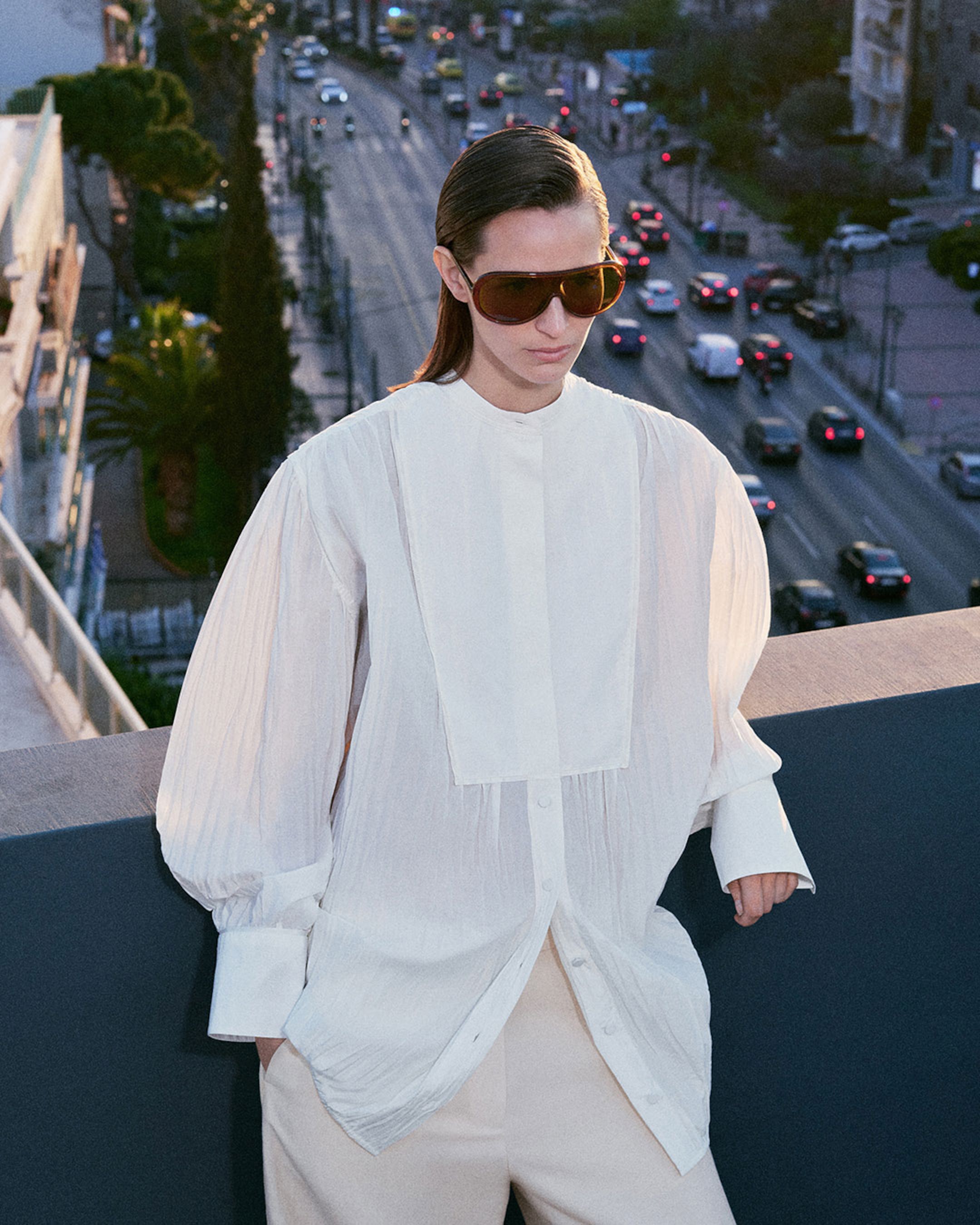 Person wearing a white shirt and sunglasses stands on a balcony overlooking a busy city street filled with cars.