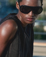 Placeholder for: Person wearing large, dark sunglasses and a sleeveless black top, gazing intently forward with a blurred outdoor background.