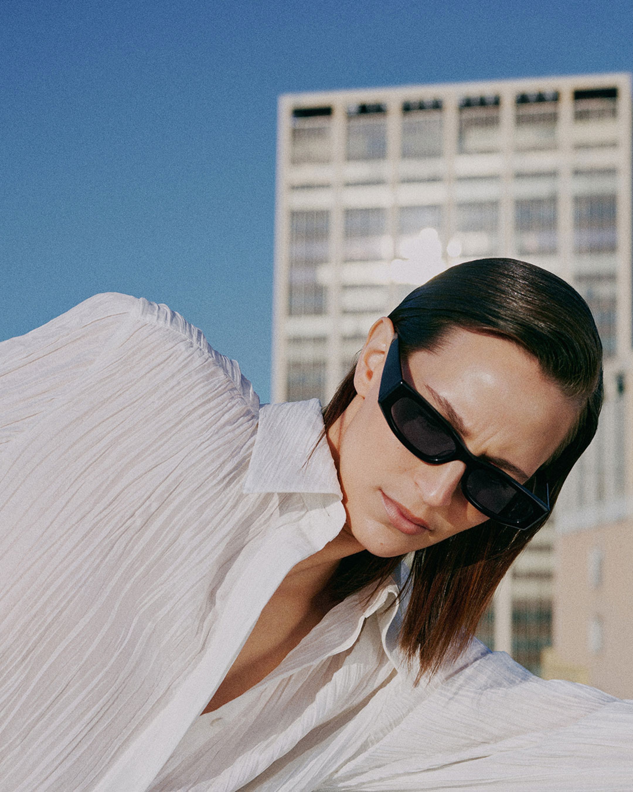 A person wearing dark sunglasses and a white textured blouse poses outdoors with a building in the background under a clear blue sky.