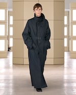 Placeholder for: Model in a long, dark trench coat with high collar and belt, walking indoors with a neutral expression. Beige panel background.