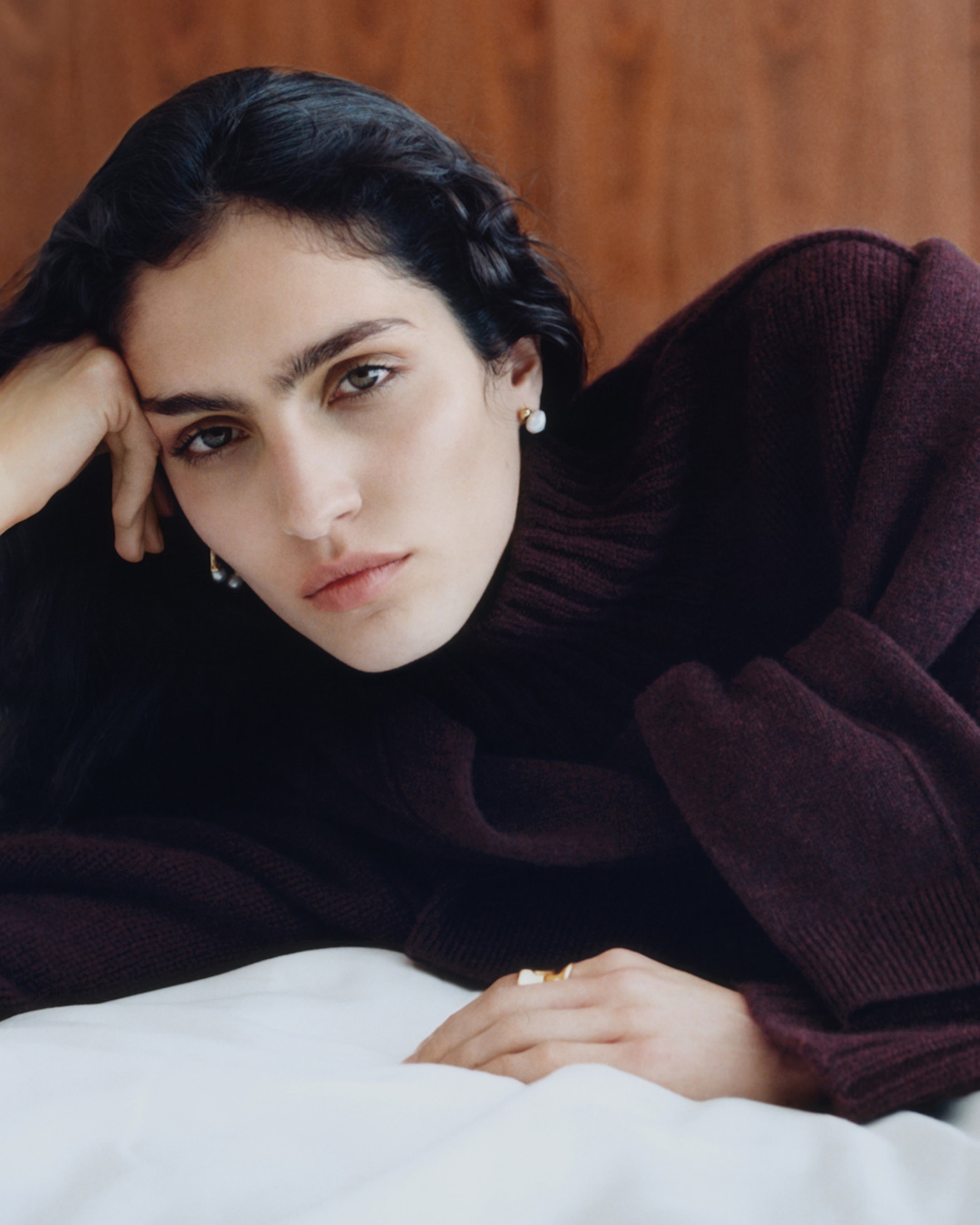 Person with dark hair wearing a burgundy turtleneck sweater, resting their head on their hand, with a neutral expression.