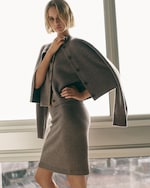 Placeholder for: Woman in a gray cardigan and skirt poses confidently by a window, with natural light illuminating the scene.