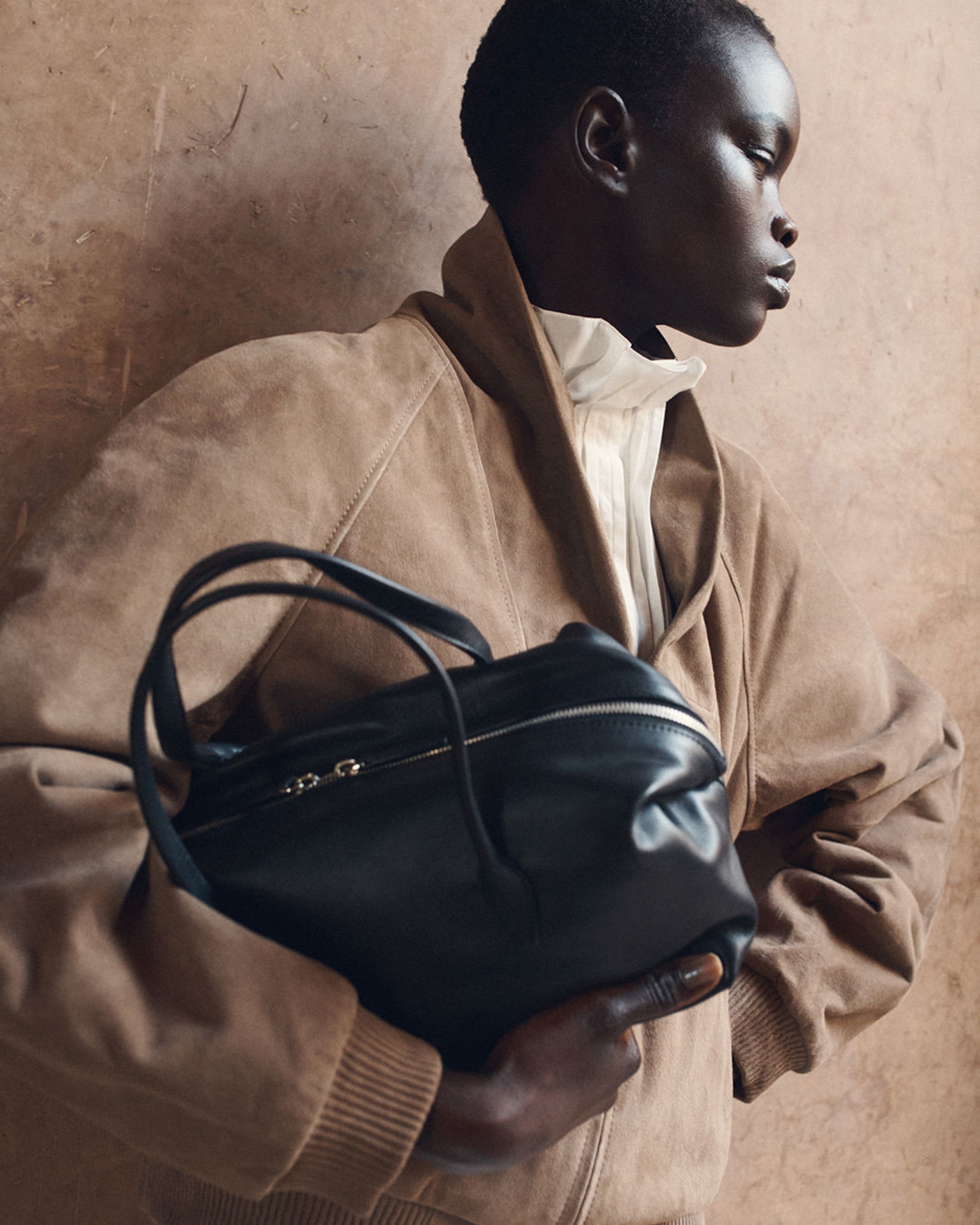 A person standing sideways, wearing a tan jacket and holding a black handbag, set against a textured beige wall.