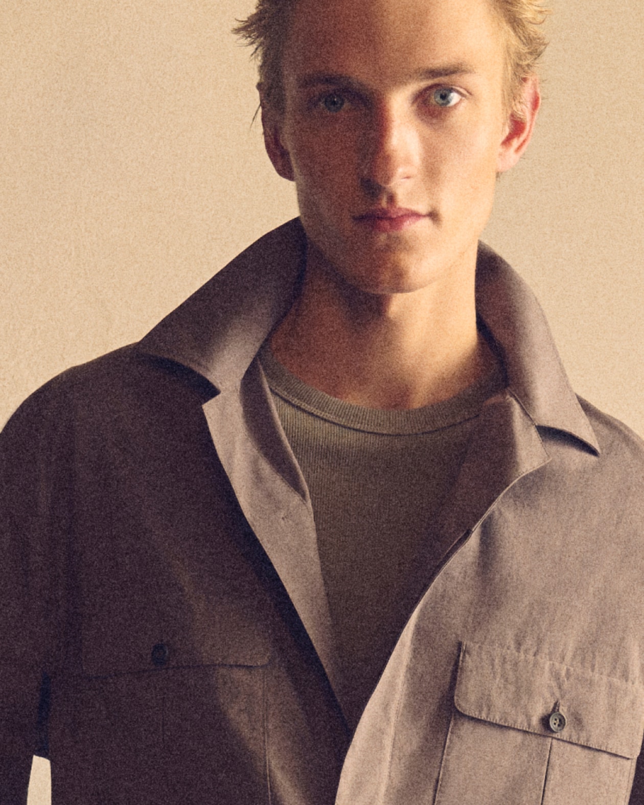 Close-up portrait of a young man in a collared jacket and knit shirt against a beige background, neutral expression.