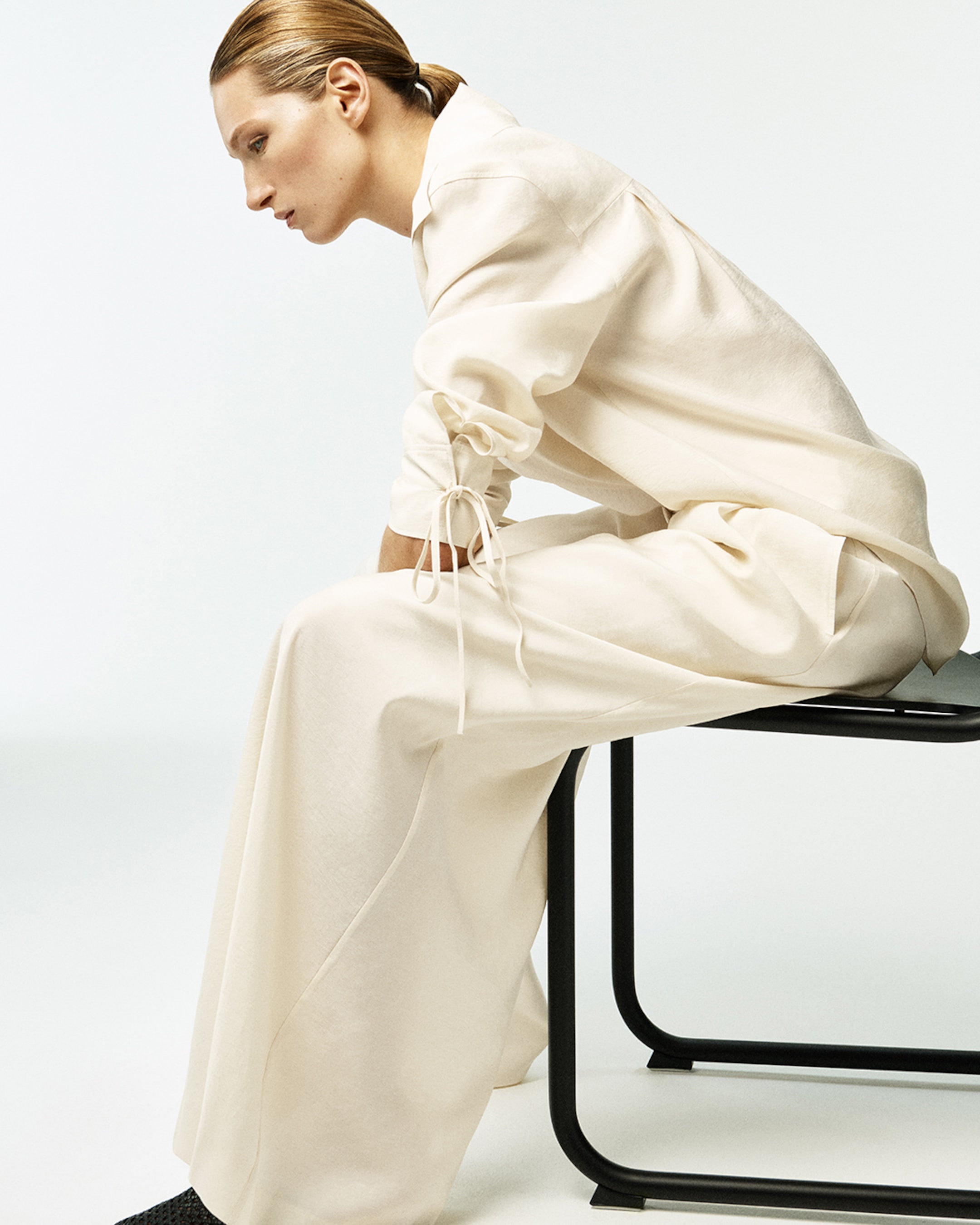 Person sitting sideways on a chair wearing a cream-colored outfit with long sleeves and pants, against a plain background.