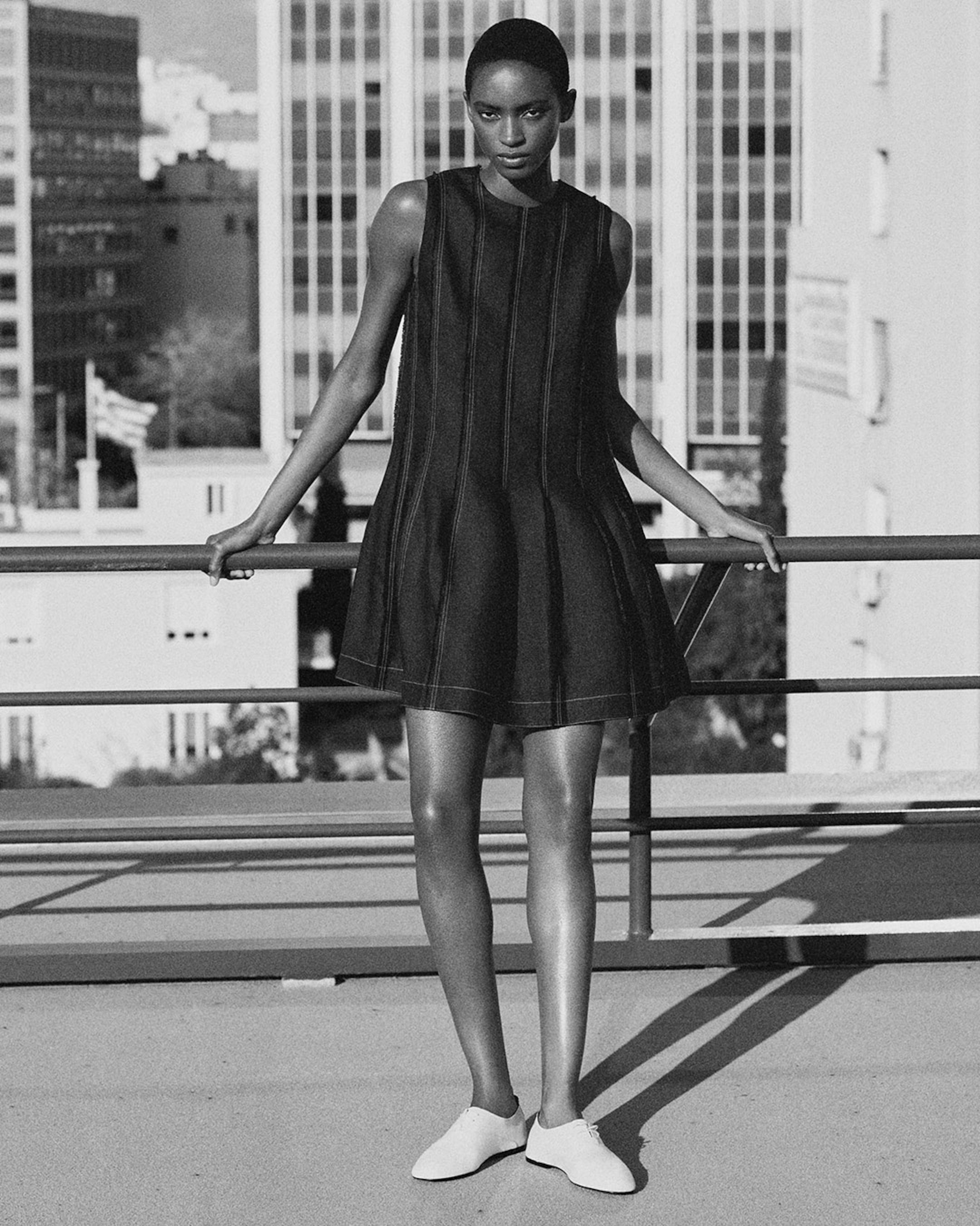 A woman in a sleeveless black dress and white shoes stands confidently on a rooftop. The city skyline is visible in the background.
