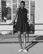 Placeholder for: A woman in a sleeveless black dress and white shoes stands confidently on a rooftop. The city skyline is visible in the background.