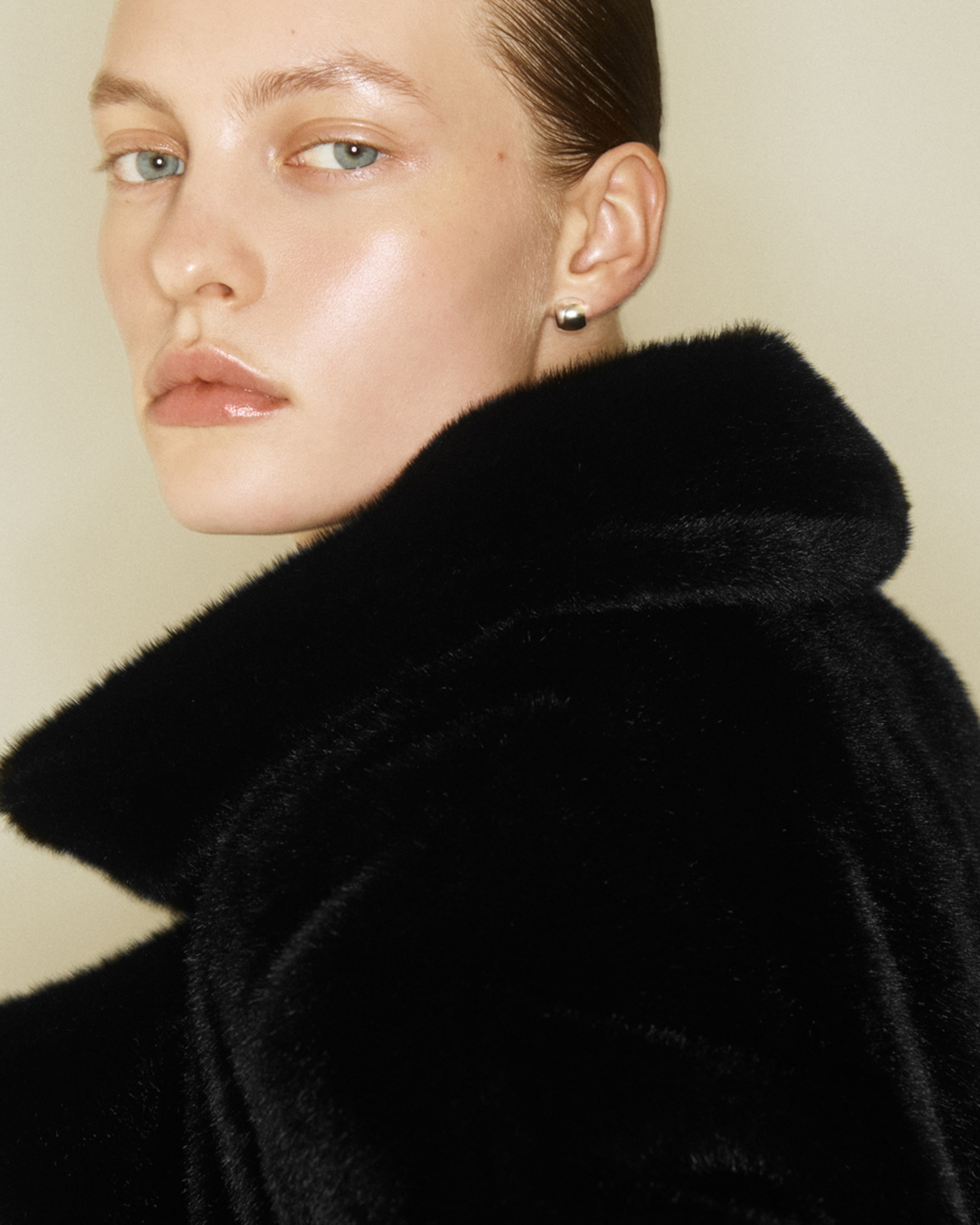 Close-up of a person with slicked-back hair, pearl earring, and black fur collar, gazing toward the camera.