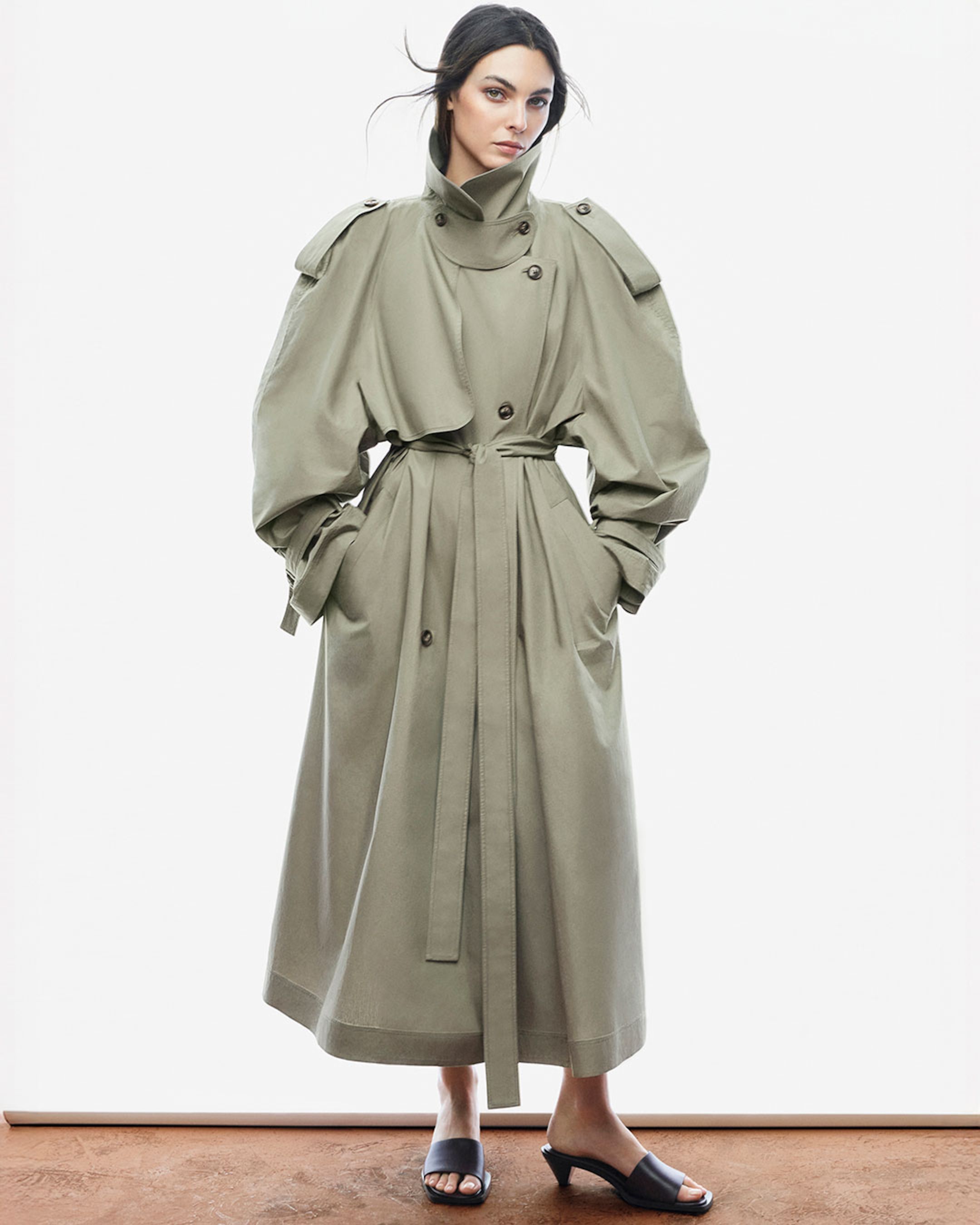 Woman in a long, olive green trench coat with a high collar and black sandals, standing against a plain white background.