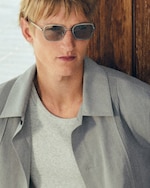 Placeholder for: Man wearing sunglasses and a light gray shirt, leaning against a wooden wall, with a neutral expression and short blond hair.