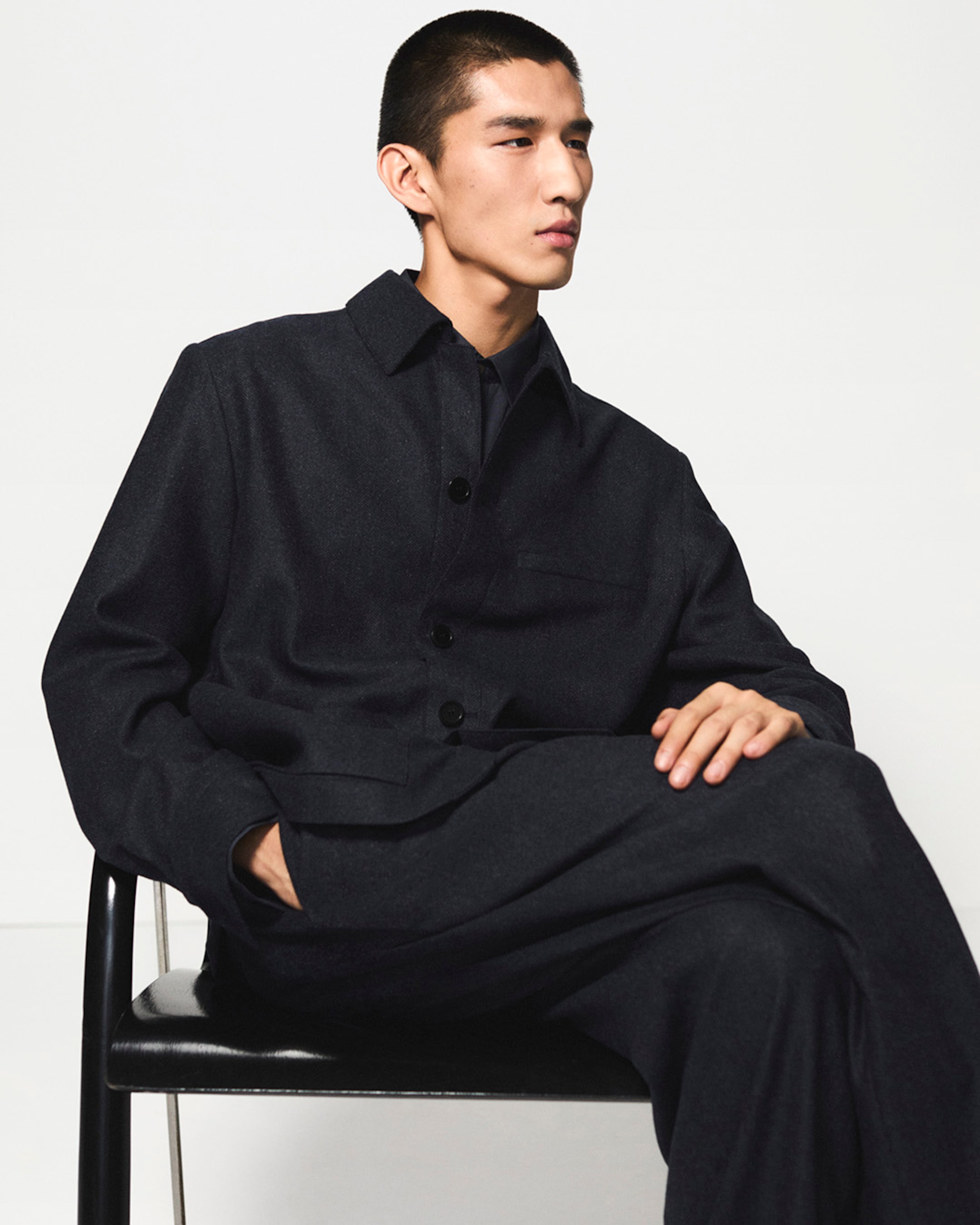 Man seated in a dark buttoned-up outfit, looking to the side against a plain background.