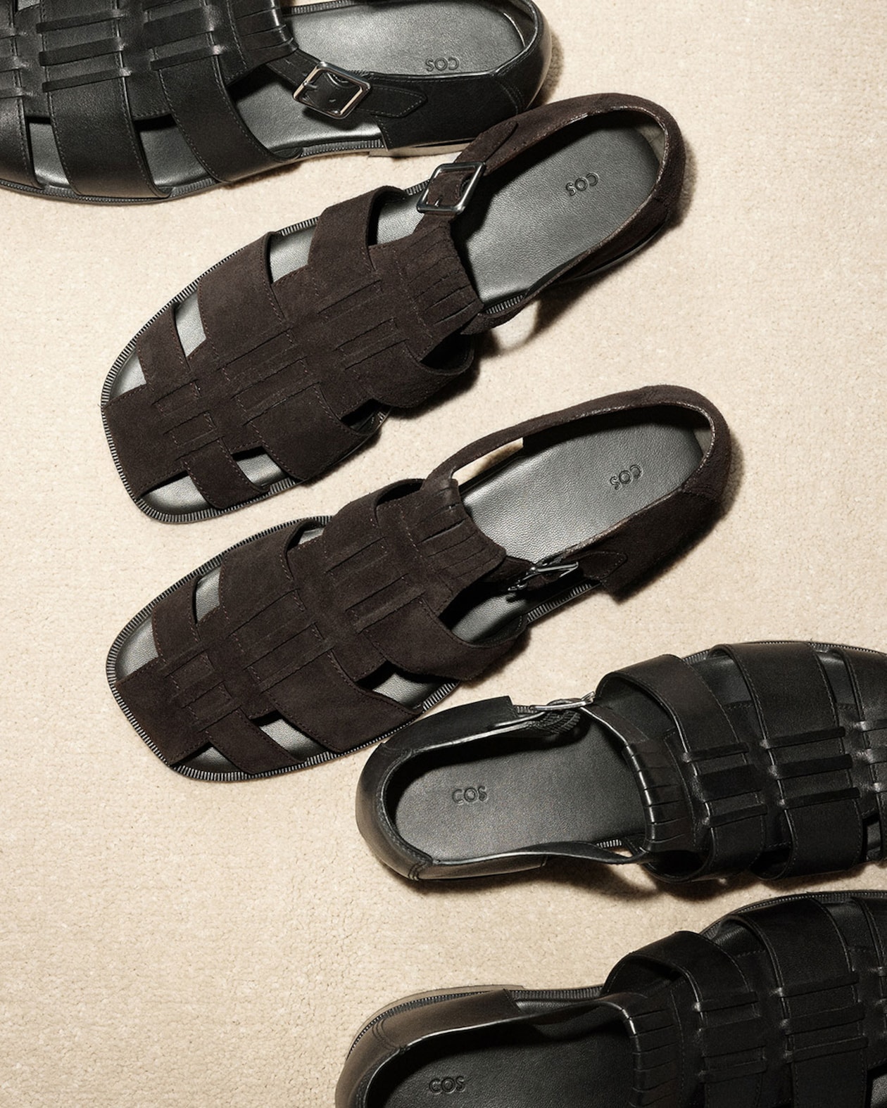 Four pairs of black woven leather sandals with buckles, arranged on a beige surface, casting soft shadows.