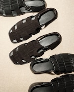 Placeholder for: Four pairs of black woven leather sandals with buckles, arranged on a beige surface, casting soft shadows.