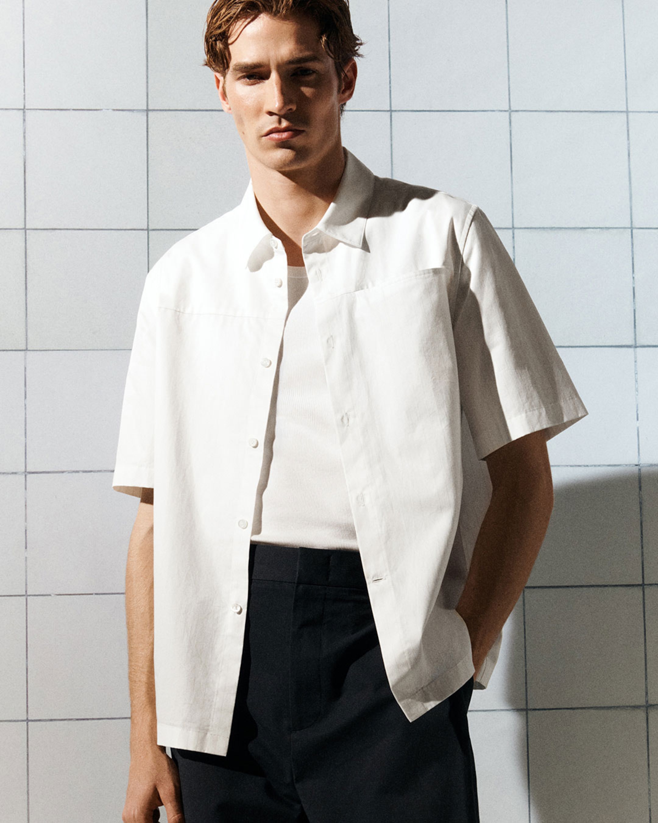 Man in a white shirt and black pants stands against a tiled wall. Hands in pocket, he gazes forward under bright lighting.