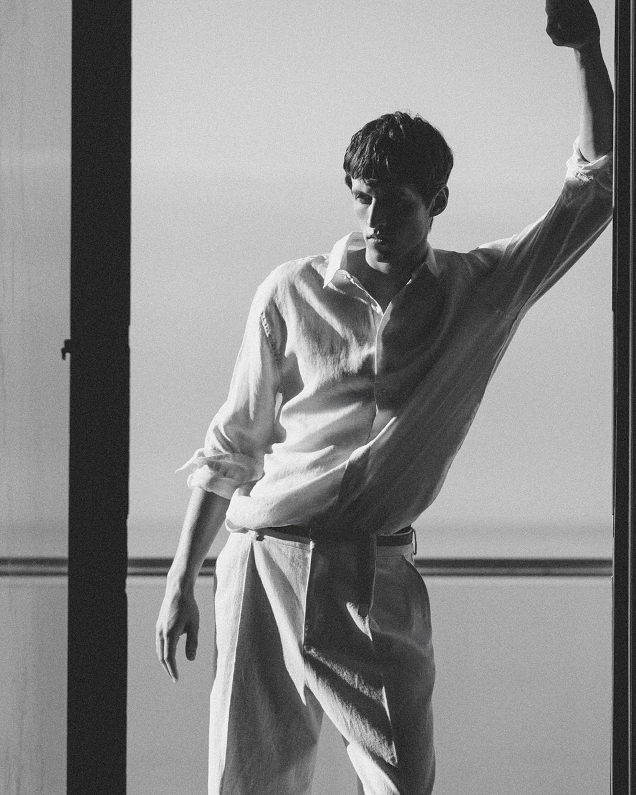 A black-and-white editorial photograph of a man in a relaxed stance, leaning one arm against a doorway frame overlooking the sea. He is wearing a breathable white linen long-sleeved shirt with the sleeves rolled up and matching light-colored tailored trousers.