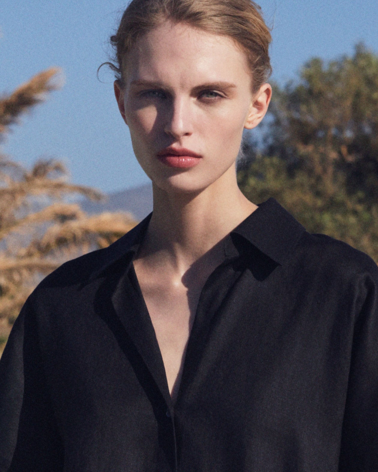 Person with light hair wearing a black shirt stands outdoors, with trees and a clear blue sky in the background.