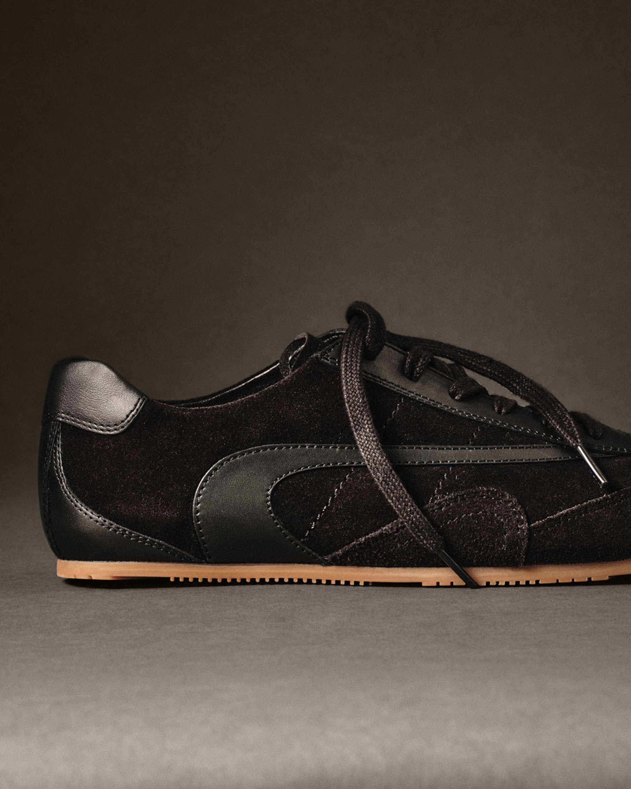 Black suede sneaker with black leather accents and brown sole, displayed on a gray surface against a dark background.
