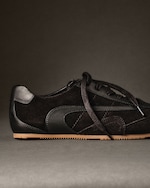 Placeholder for: Black suede sneaker with black leather accents and brown sole, displayed on a gray surface against a dark background.