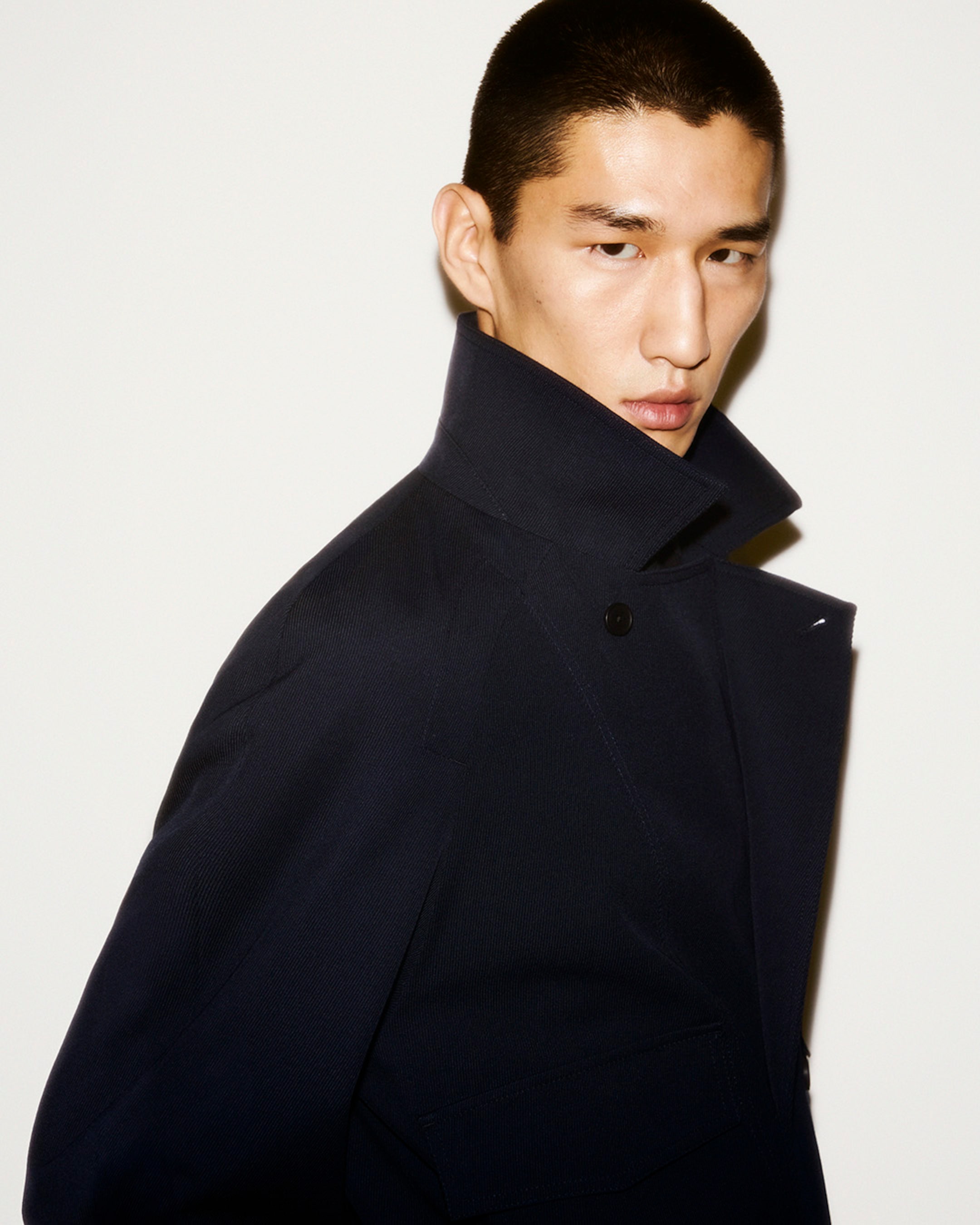Male model standing against a white background, wearing a dark high-collared jacket, looking to the side with a serious expression.