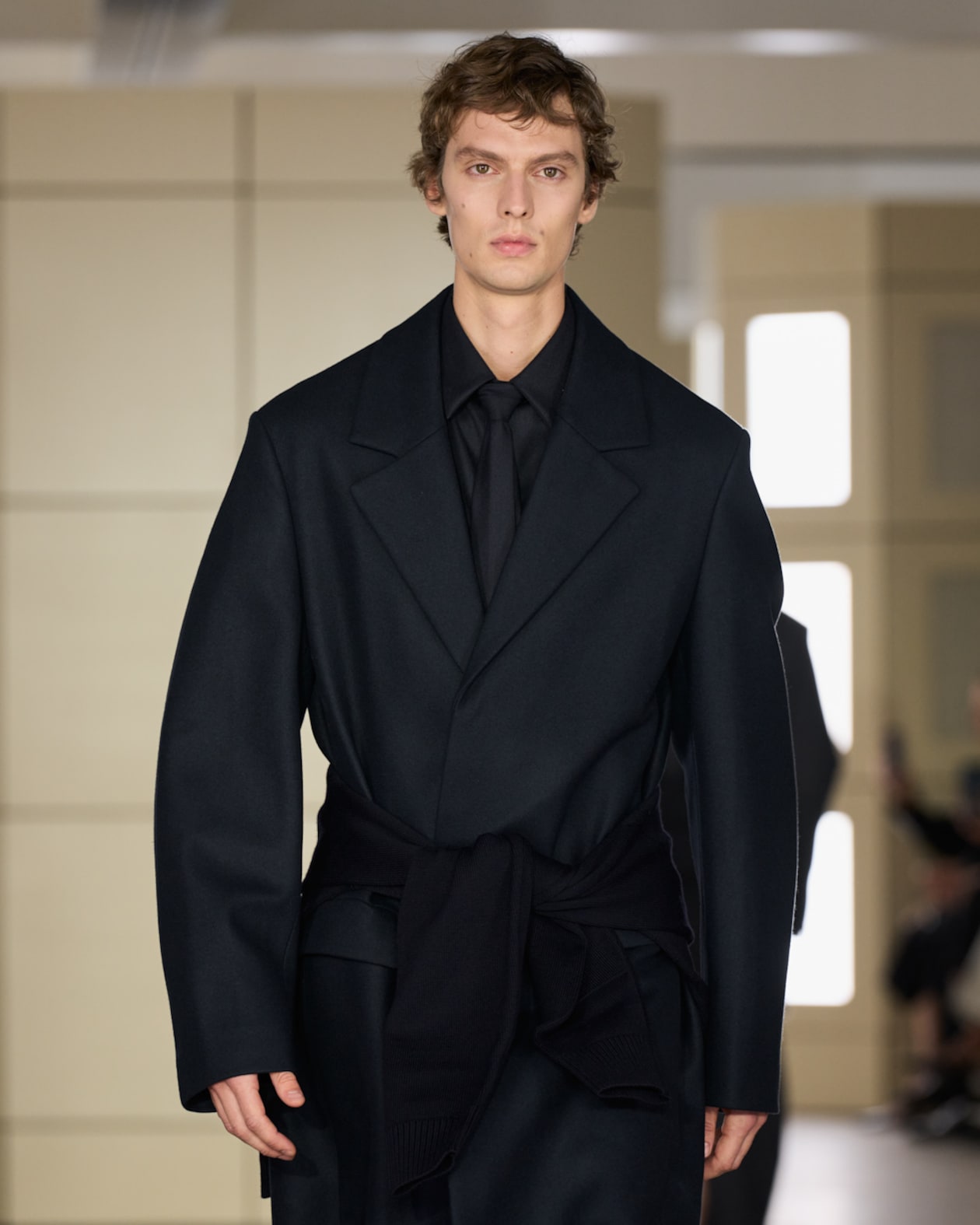 A person wearing a dark, oversized coat and a matching formal shirt and tie walks the runway in a modern, well-lit setting.