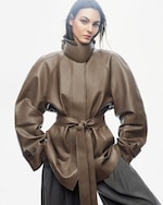 Placeholder for: A person wearing a high-collared, belted brown leather coat with wide sleeves and gray trousers, standing against a plain background.
