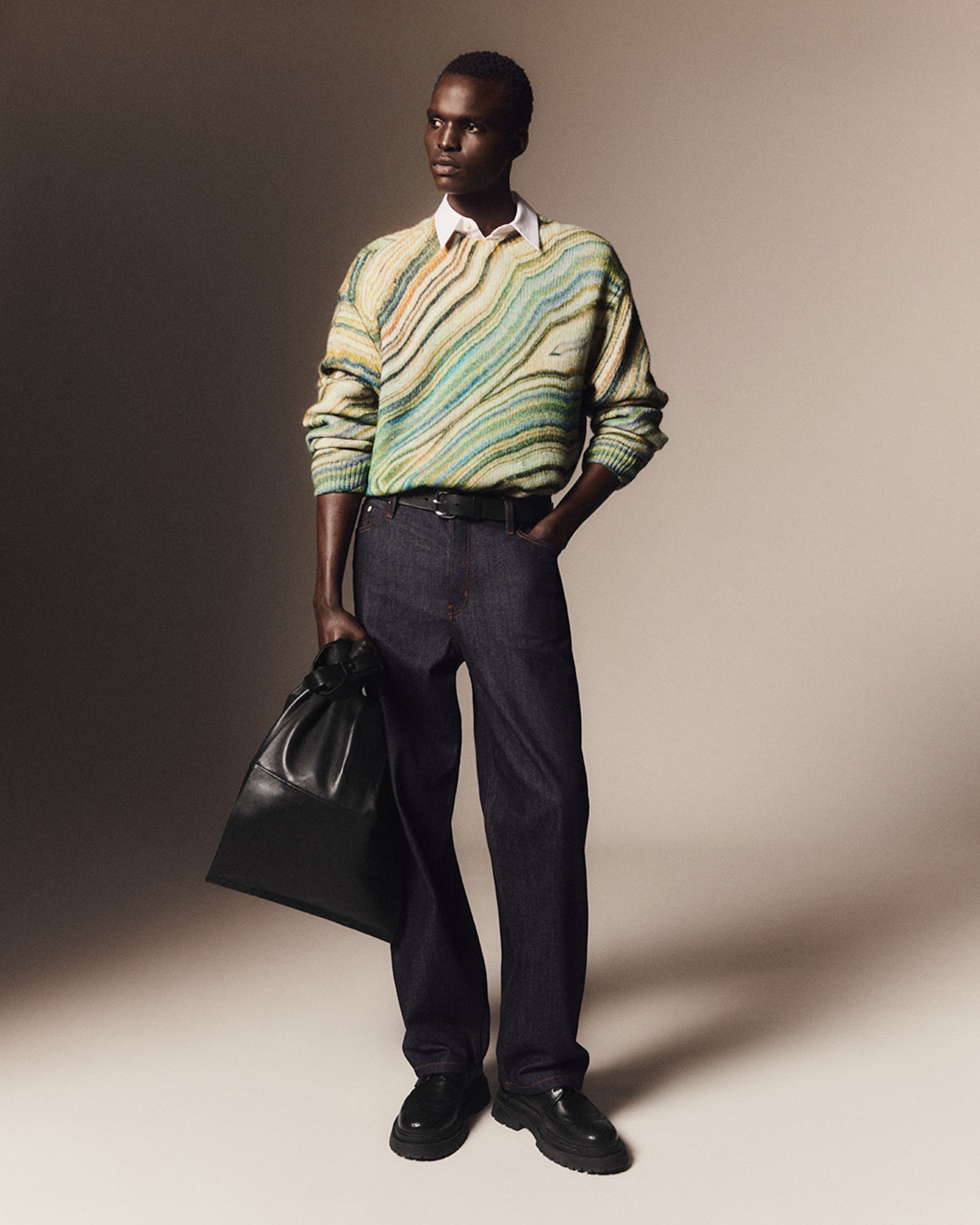 A person stands confidently wearing a green and beige patterned sweater, dark jeans, and holding a black bag against a neutral background.