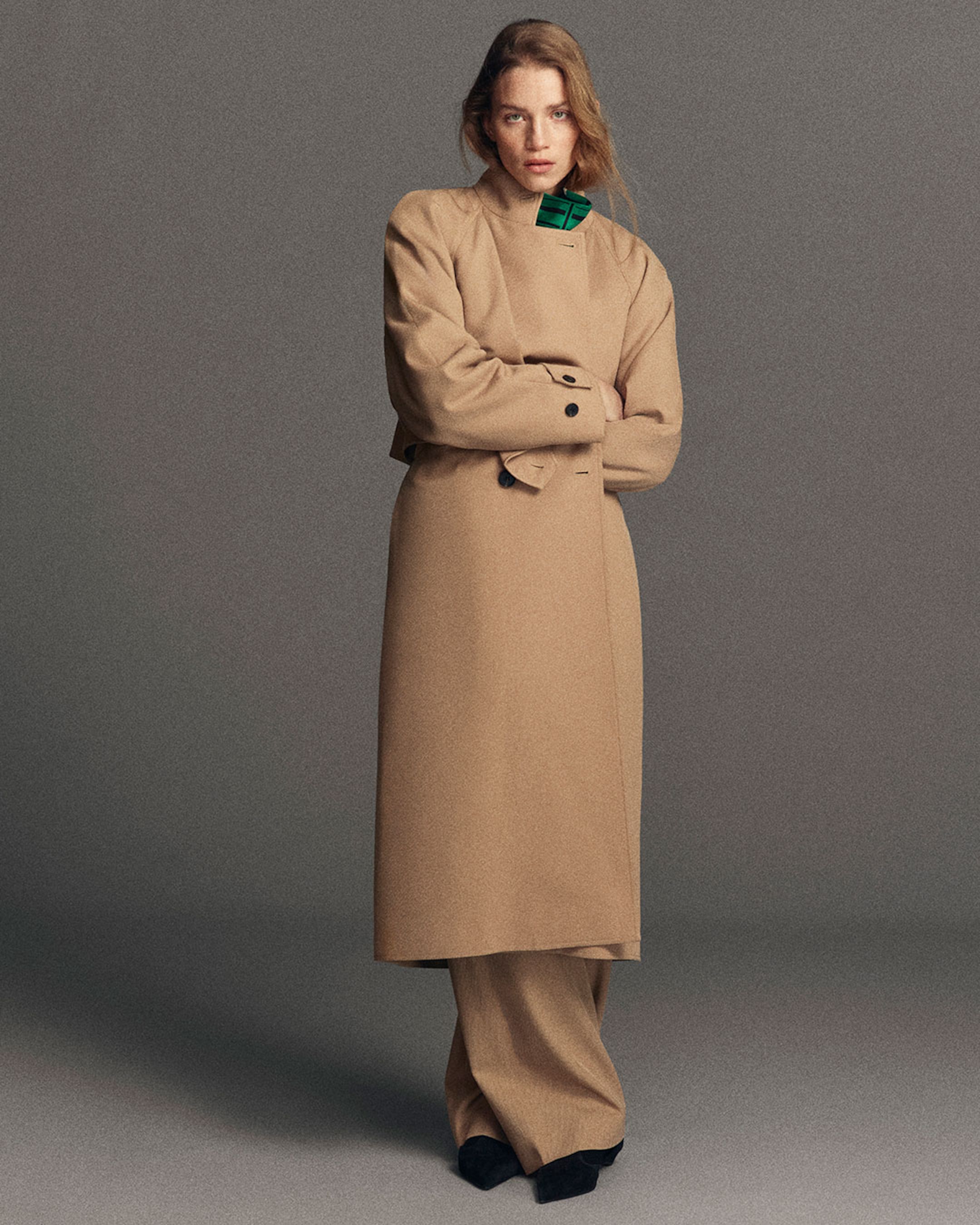 Person wearing a tan coat with black buttons and a green collar, standing with arms crossed against a gray background.