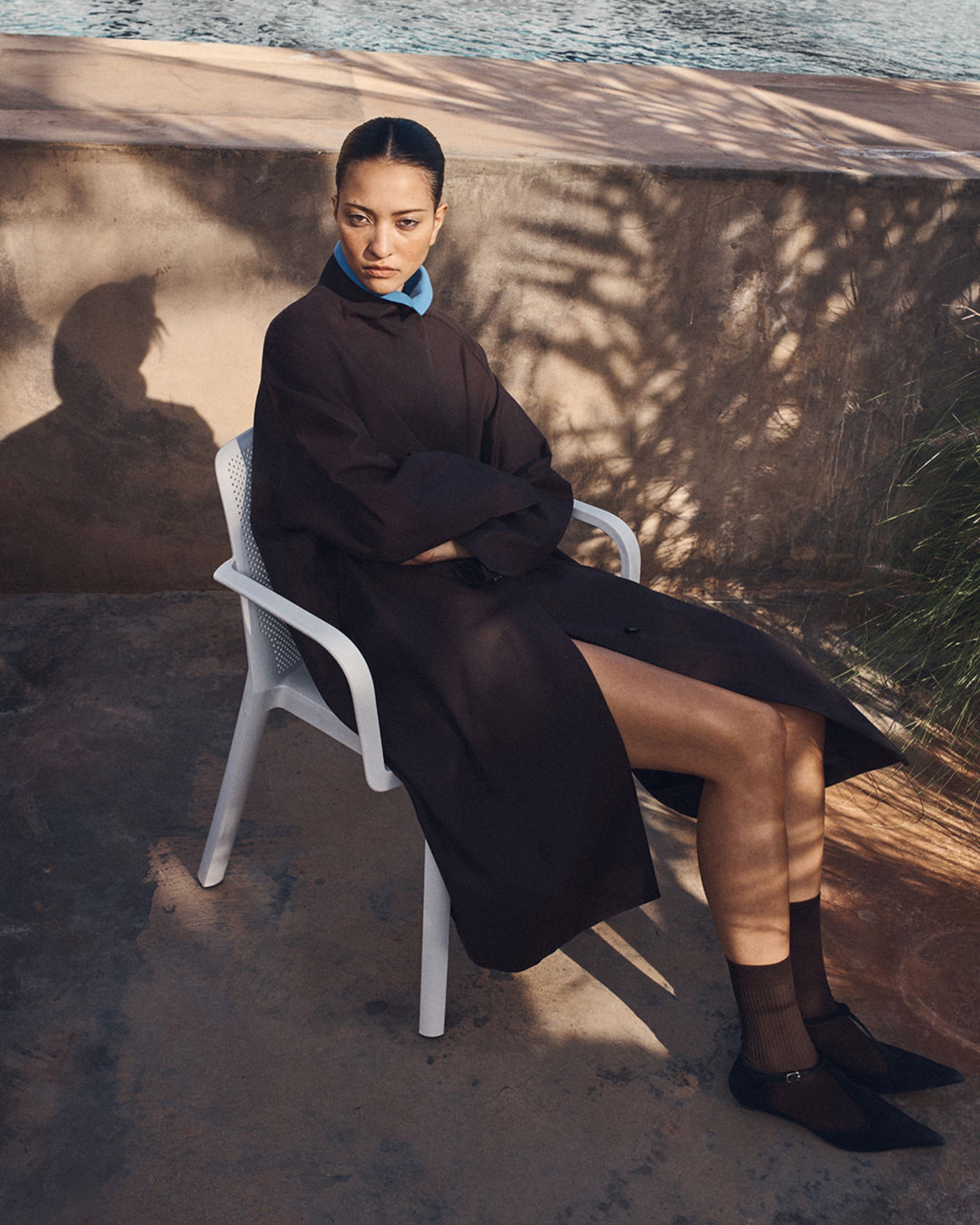 Person sitting on a white chair, wearing a dark coat with blue collar, in a sunlit outdoor setting with a stone wall and shadow patterns visible.