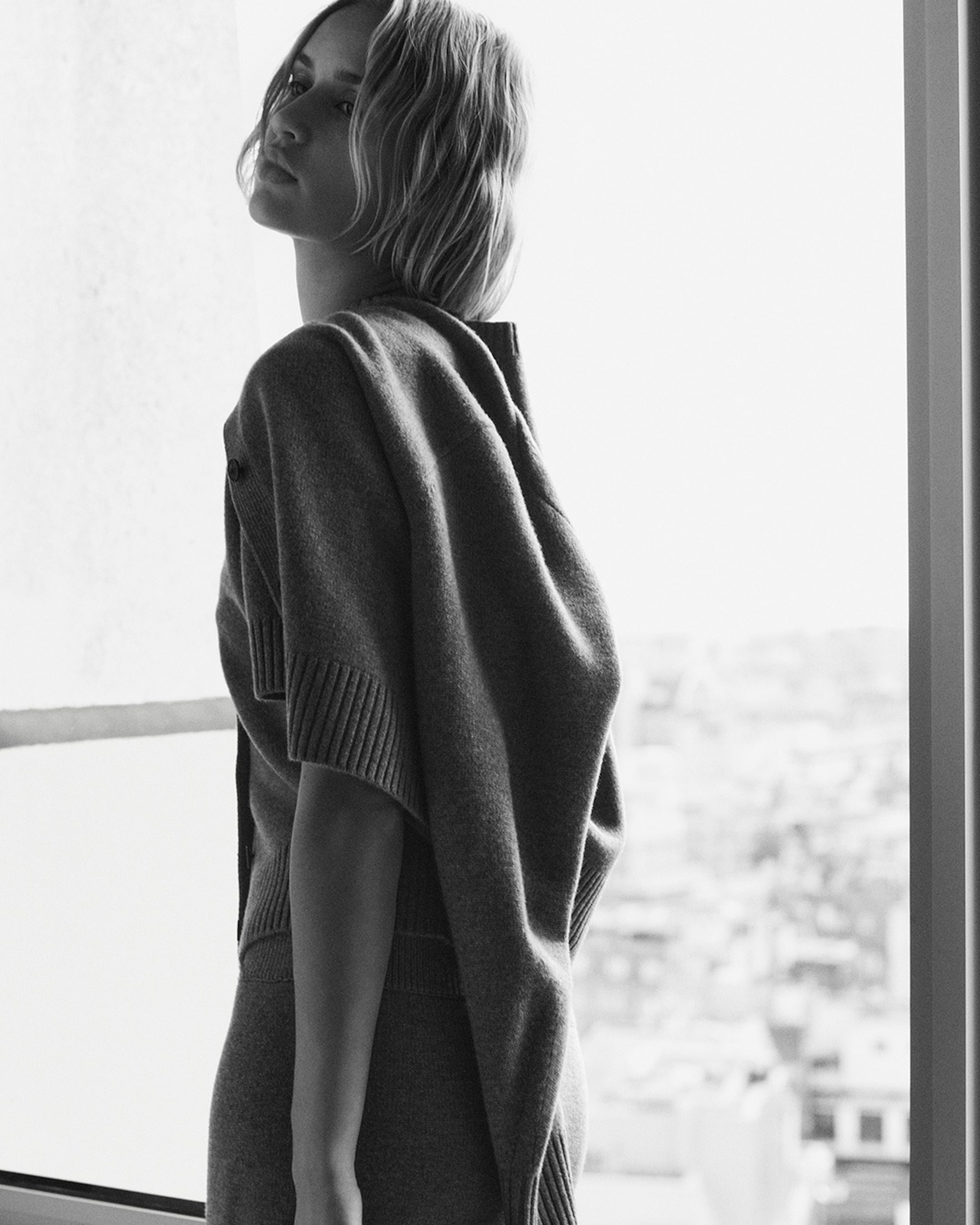 Black and white image of a woman standing by a window, wearing a sweater draped over her shoulders, with an urban landscape in the background.