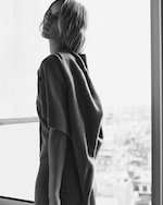 Placeholder for: Black and white image of a woman standing by a window, wearing a sweater draped over her shoulders, with an urban landscape in the background.