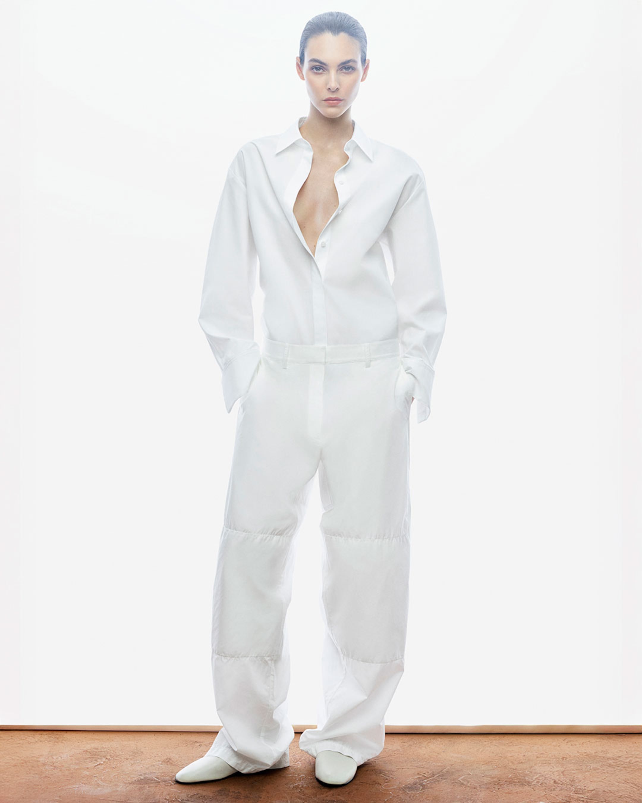A person wearing an open white shirt and loose white pants, standing against a minimalistic white background with a serious expression.
