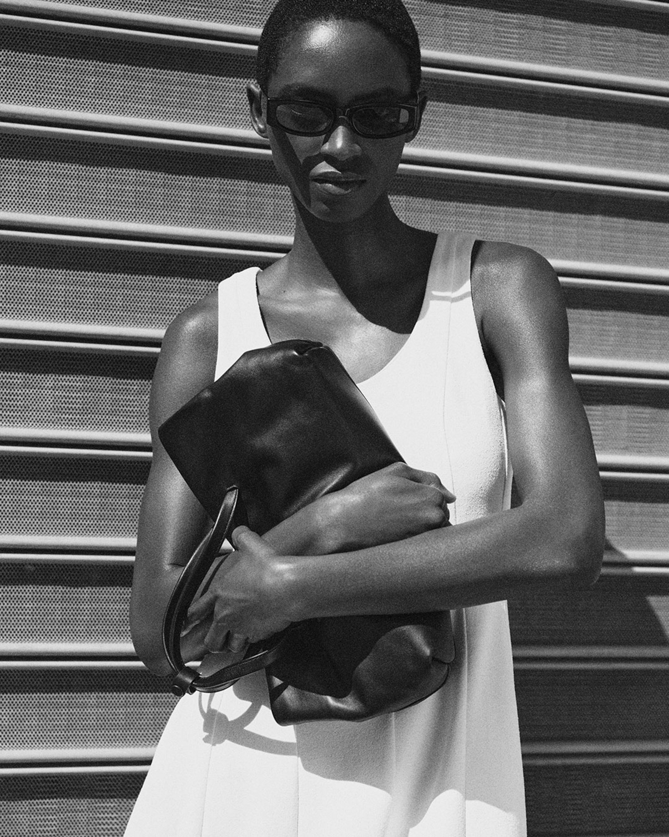 Black and white image of a woman in sunglasses, wearing a sleeveless dress, holding a leather bag, standing in front of a metal shutter.