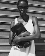 Placeholder for: Black and white image of a woman in sunglasses, wearing a sleeveless dress, holding a leather bag, standing in front of a metal shutter.