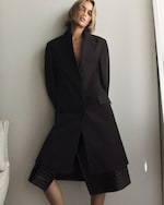 Placeholder for: Model wearing a long, dark coat, leaning against a white wall with arms behind their back, in a relaxed pose.