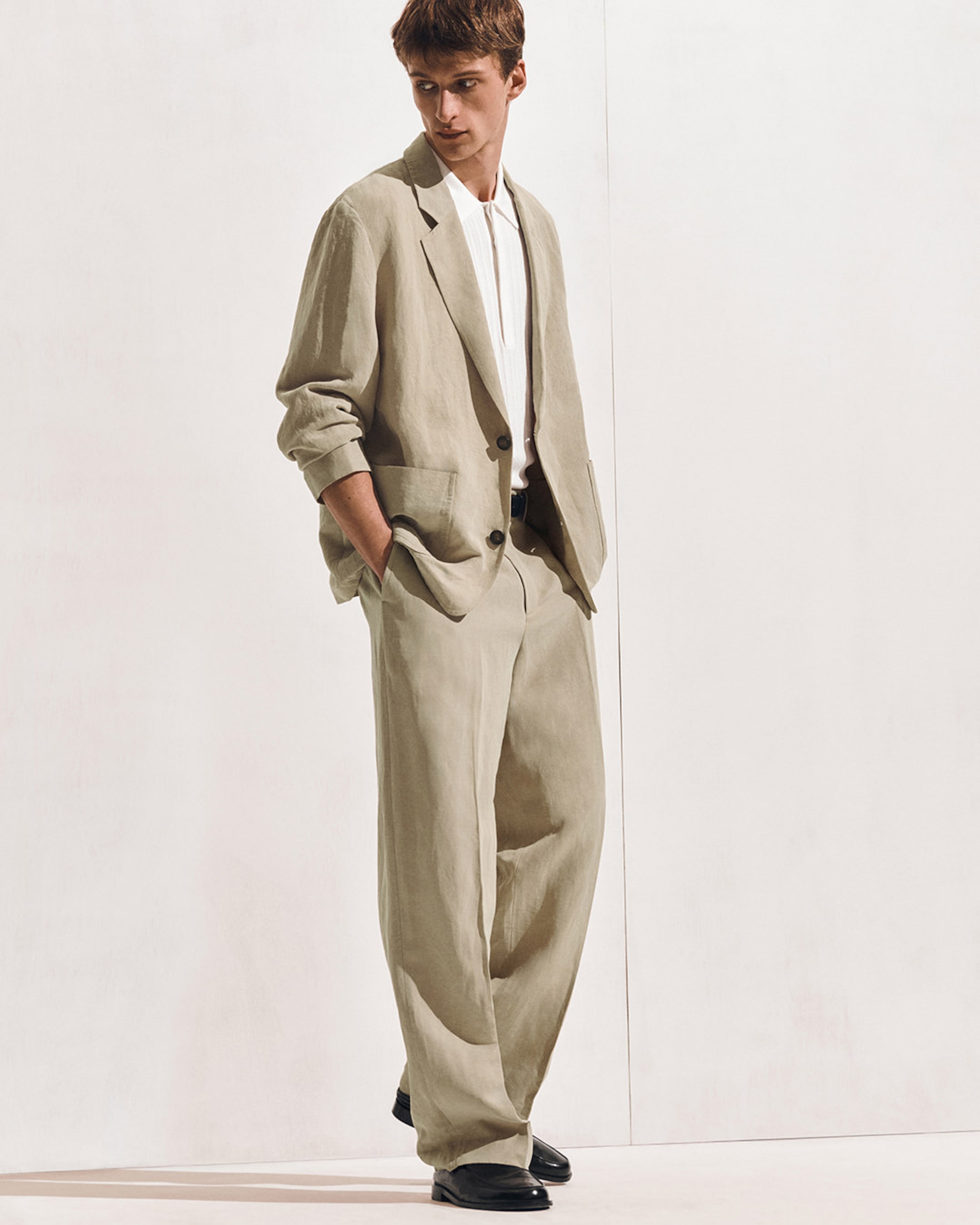 Man in a beige suit with hands in pockets, wearing a white shirt and black shoes, standing against a plain light background.