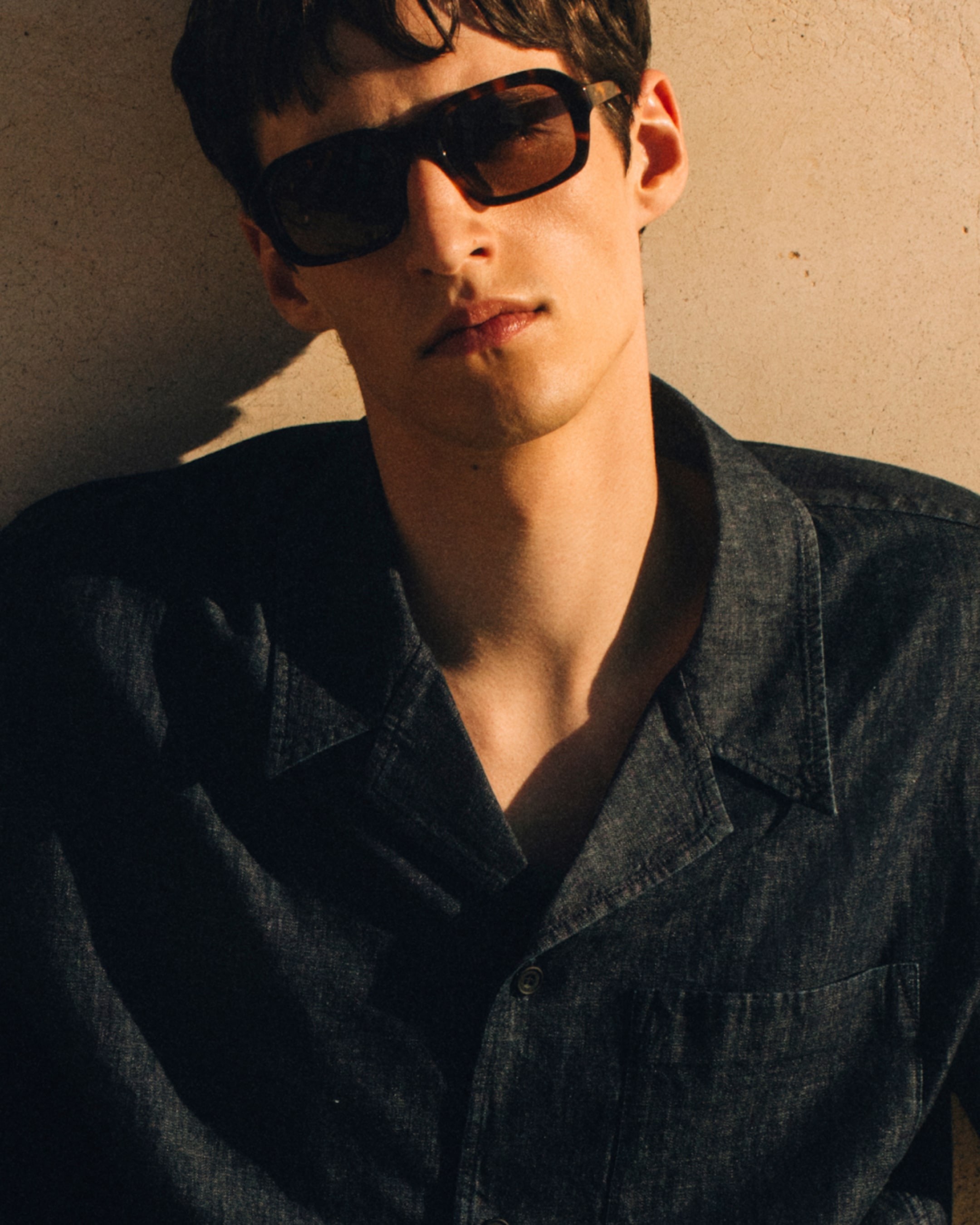 Person wearing sunglasses and a dark button-up shirt, standing against a sunlit beige wall, casting a shadow.