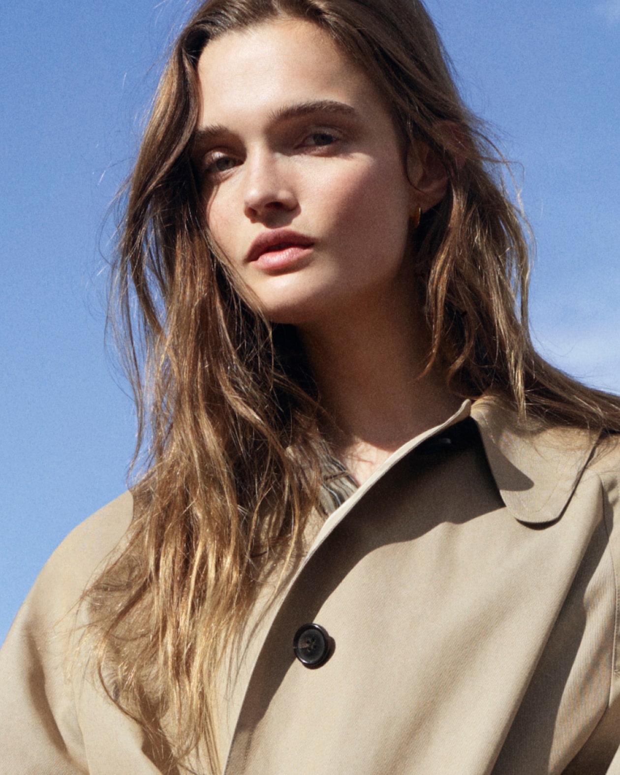 A woman with long brown hair wearing a beige trench coat stands against a clear blue sky.