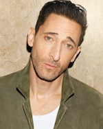 Placeholder for: A person with short dark hair and a light beard wearing a green jacket over a white shirt, standing against a textured beige background.