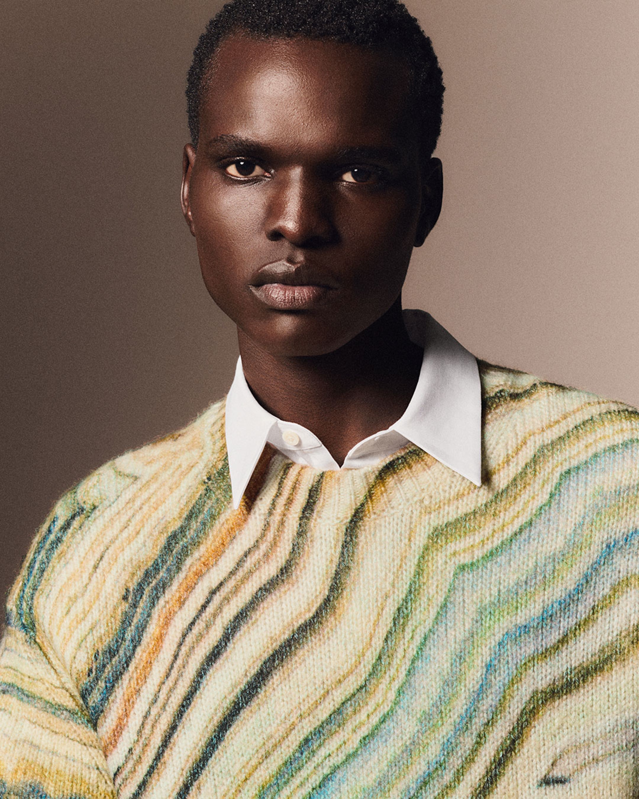 Person wearing a multicolored, wavy-patterned sweater over a white collared shirt, looking directly at the camera against a neutral background.