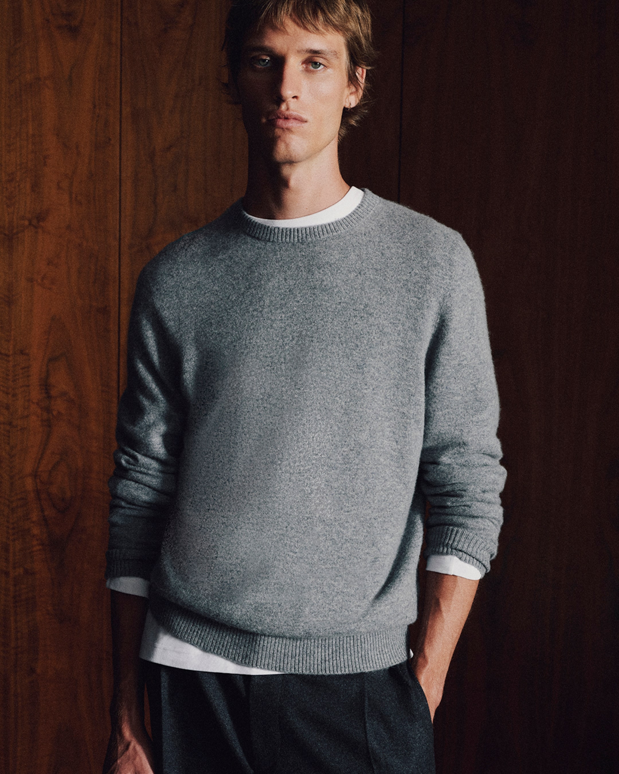 Man in a gray sweater and white shirt stands against a wooden wall, with hands in pockets, gazing at the camera.